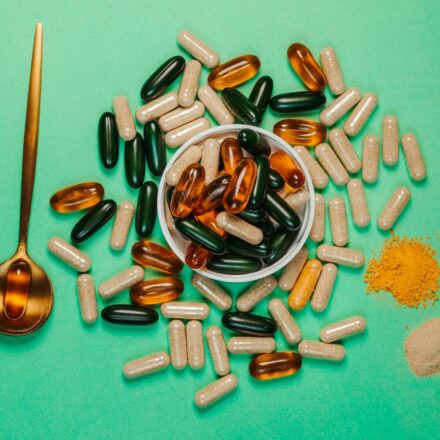 Assorted dietary supplement capsules and softgels in a bowl with turmeric powder and a gold spoon on green background.